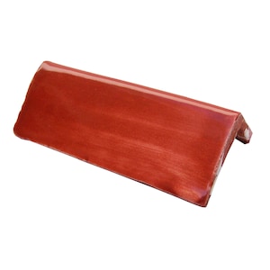 May include: A single, angled terracotta-colored roof tile. The tile has a glossy finish and a slightly curved shape. The corner of the tile is visible, showing its thickness and construction. The tile is designed for roofing applications.