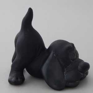May include: A black ceramic figurine of a dog in a playful pose with its tail up in the air. The dog has a cartoonish appearance with large ears and a wide, open mouth.