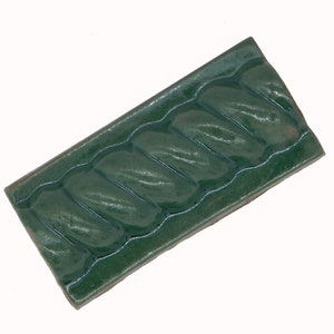 May include: A rectangular, dark green ceramic tile with a glossy finish. The tile features a repeating, raised pattern of curved, leaf-like shapes. The edges are slightly rounded, and the tile is set against a white background.