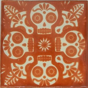 May include: A terracotta tile with a white design of four stylized skulls with floral accents. The skulls have large eye sockets and a small mouth.