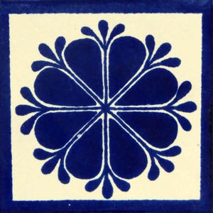 May include: A blue and white ceramic tile with a decorative floral design. The tile features a stylized flower with eight petals, each with a rounded tip. The flower is outlined in white and filled in with a deep blue color.