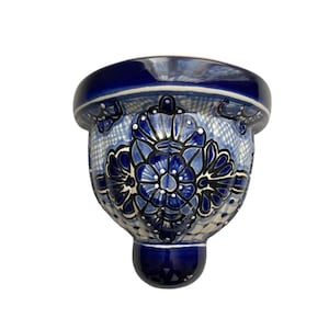 May include: A hand-painted ceramic wall pocket in a vibrant blue and white floral design. The decorative piece features a rounded top, a detailed central motif, and a small, rounded base. Ideal for displaying small plants or decorative items.