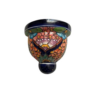 May include: A hand-painted ceramic wall pocket in a vibrant floral design. The piece features a dark blue rim and base, with a colorful pattern of flowers in orange, pink, and blue. The wall pocket is designed to hold small plants or decorative items.