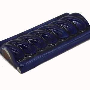 May include: A rectangular, cobalt blue ceramic tile with a braided rope design. The tile has a glossy finish and is raised in the center, with a slightly raised border. The edges show a glimpse of white and brown.