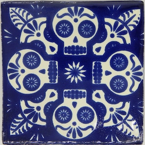 May include: A blue ceramic tile with a white design of four stylized skulls with floral accents. The skulls have large eyes and a simple nose and mouth.