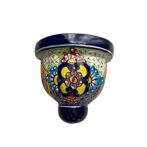 May include: A hand-painted ceramic wall planter with a vibrant floral design. The planter features a dark blue rim and base, with a colorful pattern of flowers in yellow, blue, and purple. The planter is approximately 6 inches in height.