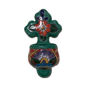 May include: A green ceramic wall pocket with a cross-shaped top and a rounded base. The top features a red and white flower design, while the base has a floral pattern with purple, blue, and yellow accents. The wall pocket is designed for decorative purposes.