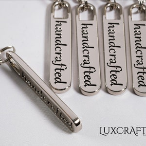 Puede incluir: Etiquetas de metal plateado con la palabra "handcrafted" grabada en ellas. Las etiquetas est&aacute;n unidas a una cadena de metal plateado con un cierre de langosta.