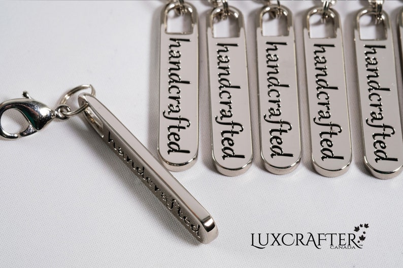 Puede incluir: Etiquetas de metal plateado con la palabra "handcrafted" grabada en ellas. Las etiquetas est&aacute;n unidas a una cadena de metal plateado con un cierre de langosta.
