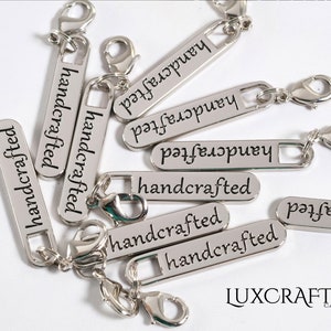 Puede incluir: Primer plano de etiquetas de metal plateado con las palabras "handcrafted" grabadas en ellas. Las etiquetas tienen un peque&ntilde;o lazo de metal y un cierre de langosta.