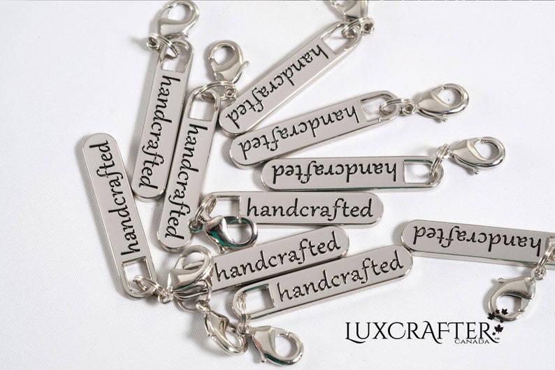 Puede incluir: Primer plano de etiquetas de metal plateado con las palabras "handcrafted" grabadas en ellas. Las etiquetas tienen un peque&ntilde;o lazo de metal y un cierre de langosta.