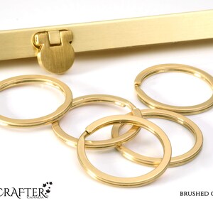 May include: Five brushed gold key rings, each with a slightly different size and shape. The key rings are arranged in a pile on a white surface. The text "BRUSHED GOLD" is visible in the lower right corner of the image.