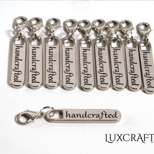 Puede incluir: Diez tiradores de cremallera plateados con la palabra "handcrafted" grabada en cada uno. Los tiradores de cremallera est&aacute;n dispuestos en una fila. Hay un tirador de cremallera adicional debajo de la fila, tambi&eacute;n con la palabra "handcrafted" grabada en &eacute;l. La palabra "LUXCRAFTER" est&aacute; impresa debajo de los tiradores de cremallera.