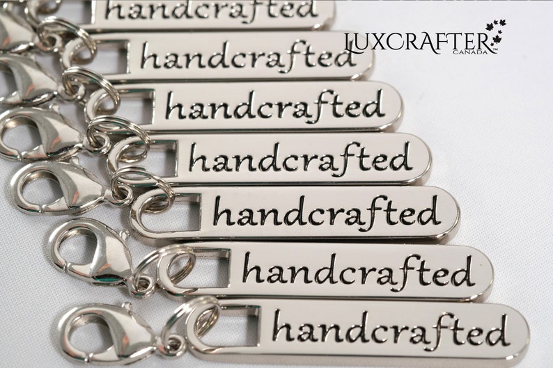 Puede incluir: Primer plano de siete tiradores de cremallera de metal plateado con la palabra "handcrafted" grabada en cada uno. Los tiradores de cremallera est&aacute;n dispuestos en una pila, con el tirador superior en la parte superior de la imagen y el tirador inferior en la parte inferior de la imagen. Los tiradores de cremallera son todos del mismo tama&ntilde;o y forma, y tienen un acabado liso y pulido.