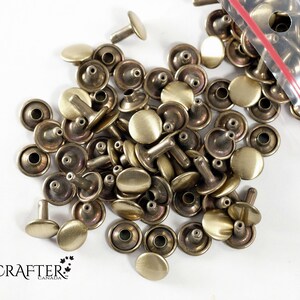 May include: A pile of gold-toned metal rivets with a round head and a post. The rivets are in a clear plastic bag with a red zipper.