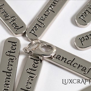 Puede incluir: Etiquetas de metal plateado con las palabras "handcrafted" grabadas en ellas. Las etiquetas est&aacute;n dispuestas de forma dispersa sobre una superficie blanca. Un cierre de metal plateado se encuentra en el centro de la imagen.