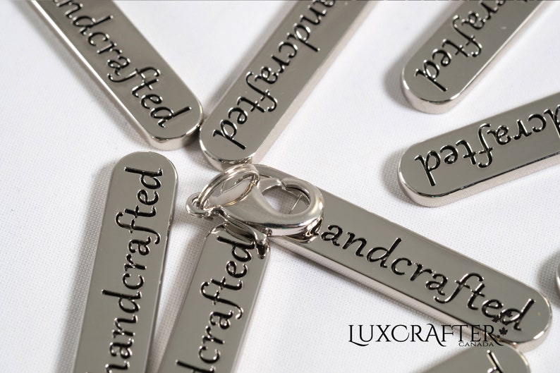 Puede incluir: Etiquetas de metal plateado con las palabras "handcrafted" grabadas en ellas. Las etiquetas est&aacute;n dispuestas de forma dispersa sobre una superficie blanca. Un cierre de metal plateado se encuentra en el centro de la imagen.