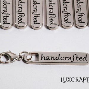 Puede incluir: Llavero de metal plateado con una etiqueta rectangular que dice "handcrafted". El llavero tiene un cierre de langosta plateado.