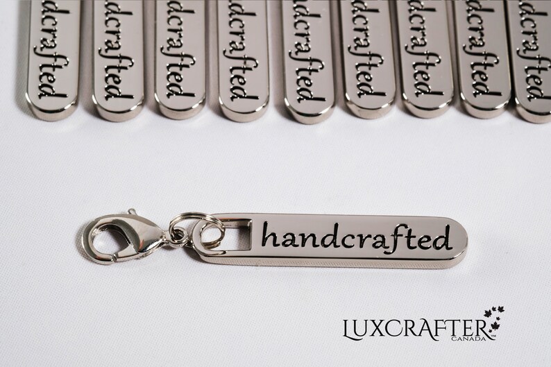 Puede incluir: Llavero de metal plateado con una etiqueta rectangular que dice "handcrafted". El llavero tiene un cierre de langosta plateado.