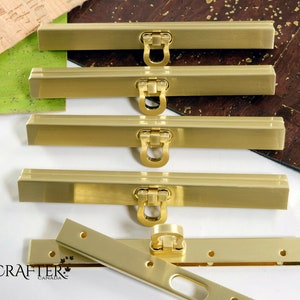 May include: A collection of gold-coloured rectangular purse clasps with ring closures. The clasps are arranged on a white surface, with cork and green material in the background. The Luxcrafter Canada logo is visible.