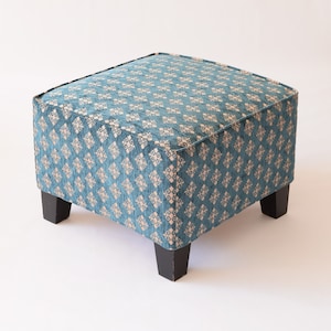 Pode incluir: Um pufe quadrado com uma capa de tecido azul turquesa com um padrão geométrico branco. O pufe tem quatro pernas de madeira pretas.