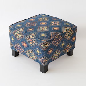 May include: A square ottoman with a blue fabric cover featuring a geometric pattern in shades of orange, yellow, and brown. The ottoman has four dark wood legs.