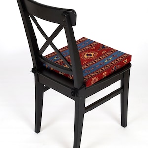 May include: Black wooden chair with a red, blue, and yellow patterned cushion. The chair has a cross-shaped back and a simple design.