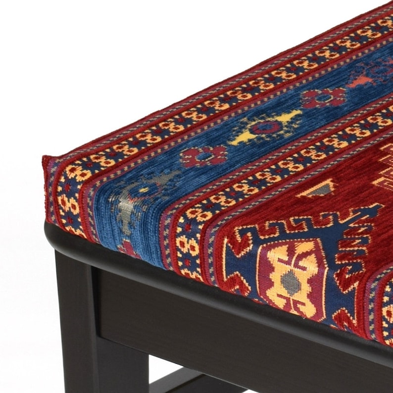 May include: A dark wood ottoman with a red, blue, and yellow patterned fabric top. The fabric has a traditional design with geometric shapes.