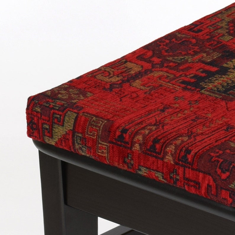 Peut inclure: Un ottoman en bois fonc&eacute; avec un dessus en tissu rouge &agrave; motifs. Le tissu pr&eacute;sente un motif traditionnel avec des accents dor&eacute;s et noirs.