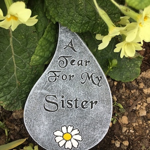 May include: A grey stone shaped like a teardrop with the text "A Tear For My Sister" and a white daisy with a yellow centre.