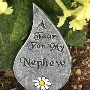 May include: A grey stone shaped like a teardrop with the words "A Tear For My Nephew" and a white daisy with a yellow centre.