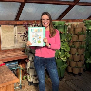 Op de afbeelding: Een vrouw in een roze trui en blauwe jeans houdt een boek vast met de titel "My Garden" in een kas. De omslag van het boek toont een kleurrijke illustratie van verschillende groenten en kruiden.