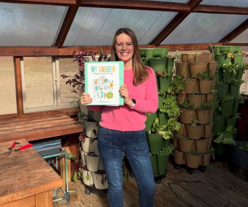 Op de afbeelding: Een vrouw in een roze trui en blauwe jeans houdt een boek vast met de titel "My Garden" in een kas. De omslag van het boek toont een kleurrijke illustratie van verschillende groenten en kruiden.