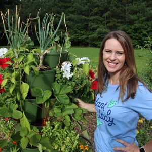 May include: A woman wearing a blue shirt with the text "Plant Lady" stands next to a vertical garden tower. The tower has four tiers with different plants growing in each tier, including red and white flowers, green leafy plants, and spring onions.