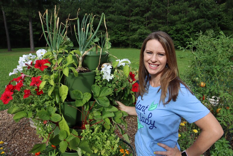 May include: A woman wearing a blue shirt with the text "Plant Lady" stands next to a vertical garden tower. The tower has four tiers with different plants growing in each tier, including red and white flowers, green leafy plants, and spring onions.