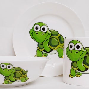 Könnte beinhalten: Ein weißes Keramik-Kindergeschirr-Set mit einem Cartoon-Schildkröten-Design. Das Set enthält eine Schüssel, einen Teller und eine Tasse.