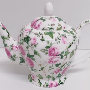 Könnte beinhalten: Eine weiße Teekanne mit einem rosa-grünen Blumenmuster. Die Teekanne hat einen Deckel und einen Henkel.
