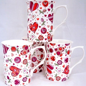 May include: Set of four white ceramic mugs with a pink and red heart pattern. The mugs have a white background with pink and red hearts, roses, and dots.