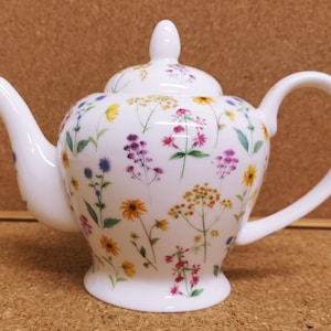 Può includere: Teiera in porcellana bianca con un motivo floreale con fiori selvatici gialli, rosa, viola e blu. La teiera ha un coperchio e un manico.
