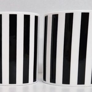 Może przedstawiać: Dwa białe ceramiczne kubki z czarnym pionowym wzorem w paski. Każdy kubek ma zakrzywiony uchwyt i cylindryczny kształt. Kubki są prezentowane na gładkim białym tle.