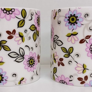 Può includere: Due tazze in ceramica bianca con un motivo floreale. Le tazze hanno uno sfondo bianco con fiori e foglie rosa, blu e verdi.