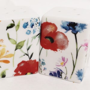 Peut inclure: Un ensemble de deux salières et poivrières en céramique blanche avec un motif floral. Les salières présentent un motif de fleurs rouges, jaunes, bleues et roses de style aquarelle sur un fond blanc.