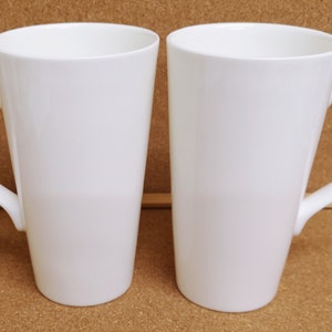 May include: Two white ceramic mugs with tall, straight sides and a handle on the right side.