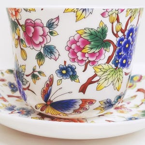 May include: A white teacup and saucer set with a floral and butterfly design. The cup and saucer feature pink, blue, yellow, and orange flowers and butterflies. The teacup is approximately 3 inches tall and 4 inches wide.