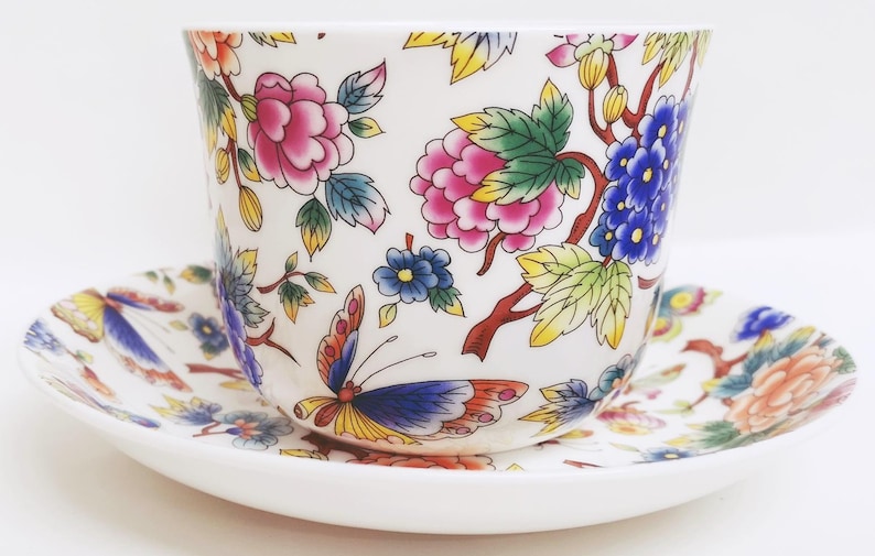 May include: A white teacup and saucer set with a floral and butterfly design. The cup and saucer feature pink, blue, yellow, and orange flowers and butterflies. The teacup is approximately 3 inches tall and 4 inches wide.