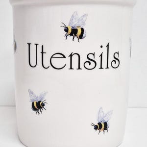 Könnte beinhalten: Weißer Keramik-Utensilienhalter mit dem Wort "Utensils" in schwarzer Schrift. Der Halter ist mit Illustrationen von fliegenden Bienen verziert. Die Bienen haben schwarz-gelbe Streifen und durchscheinende Flügel. Ein Küchendekorationsartikel.