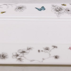 Peut inclure: Un plat de service rectangulaire blanc avec un motif floral. Le motif présente des contours noirs de fleurs, de feuilles et de papillons. Les papillons sont dans des tons de bleu, rose et vert.
