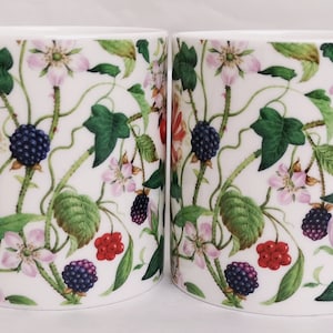 May include: Two white ceramic mugs with a botanical design. The mugs feature a repeating pattern of green leaves, pink and white flowers, red berries, and dark blue blackberries. Each mug has a white handle.