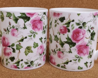 May include: Two white ceramic mugs with a pink rose floral pattern. The mugs have a white background with pink roses and green leaves.