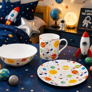 Puede incluir: Un juego de vajilla de cerámica blanca compuesto por un cuenco, una taza y un plato, decorados con un patrón colorido de planetas, estrellas y transbordadores espaciales. Los artículos están dispuestos sobre una superficie azul oscuro.